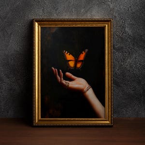 May include: A framed artwork featuring a hand with rings and a bracelet, reaching towards a vibrant orange and black butterfly against a dark background. The frame is gold-colored.