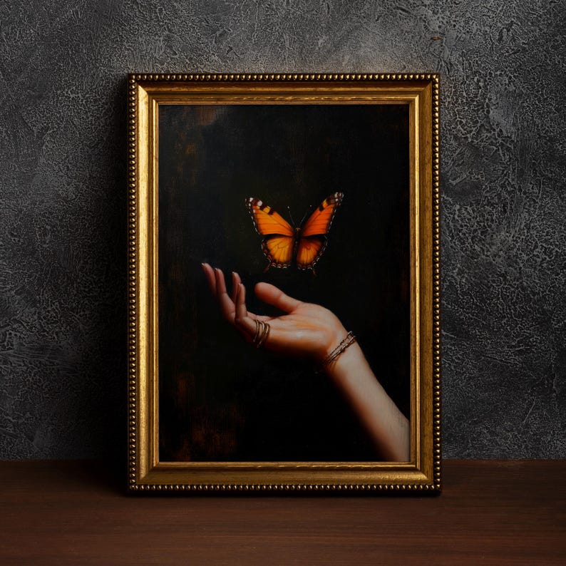 May include: A framed artwork featuring a hand with rings and a bracelet, reaching towards a vibrant orange and black butterfly against a dark background. The frame is gold-colored.