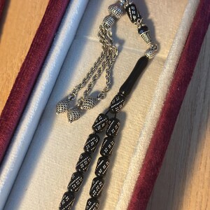 May include: A black prayer beads necklace with silver accents and a silver tassel. The beads are cylindrical and have a white pattern. The necklace is in a red velvet box.