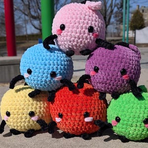 May include: Six crocheted plush toys in different colors, including pink, blue, purple, yellow, red, and green. Each toy has a round body with small black eyes and a stitched-on mouth. The toys have black crocheted legs and arms.