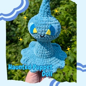 May include: A handmade, light blue crochet doll with a unique design. The doll has large, blue eyes with yellow accents, a small smile, and a flared base. The text "Haunted Puppet Doll" is displayed on the doll.