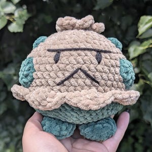 May include: A handmade crochet plush toy in the shape of a grumpy-faced creature. The toy features a tan body with green accents on the sides and bottom. Black yarn is used for the eyes and mouth. The plush is held in a hand against a blurred green background.