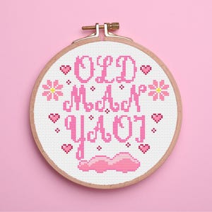 May include: A cross-stitch embroidery hoop with the words "OLD MAN YAOJ" in pink. The design includes pink hearts, flowers, and a cloud-like shape. The hoop is set against a pink background.