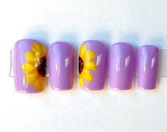 May include: Set of five lavender-colored press-on nails. Two nails feature a yellow sunflower design. The nails are rectangular in shape and are displayed on a clear adhesive strip against a white background. Nail art.