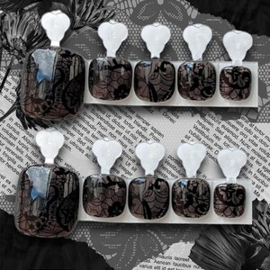 May include: Set of press-on nails with a black lace design. Each nail is a glossy dark color with a detailed lace pattern. The nails have a white tab for easy application. The background includes floral and text elements.