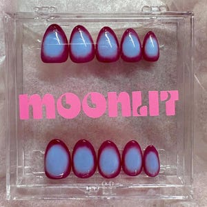 May include: A set of press-on nails in a clear acrylic case. The nails feature a gradient design with a light blue center and a dark magenta border. The word "MOONLIT" is printed in pink.