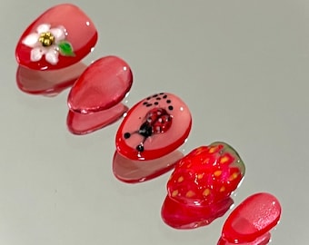 May include: Five press-on nails with a glossy red finish. One nail features a white flower with a gold center, another displays a ladybug, and a third shows a textured strawberry design. The remaining two nails are solid red with a subtle shimmer.