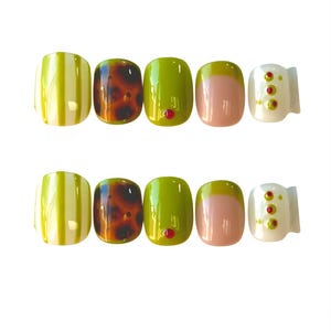 May include: A set of press-on nails in various designs. The nails feature stripes, tortoiseshell, solid olive green with a red gem, a pink and yellow gradient, and white with red and yellow accents. The nails are arranged in two rows.