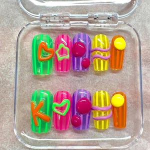 May include: A set of ten press-on nails in a clear plastic case. Each nail features vertical stripes in neon green, pink, purple, yellow, and orange. The nails are decorated with raised 3D elements including an orange letter K, green heart, purple circles, lavender wavy lines, and yellow circles.