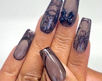 May include: A set of long, square-shaped artificial nails. The nails feature a dark brown base with a black lace overlay. One nail has a black bow accent. The design is elegant and detailed, suitable for special occasions.