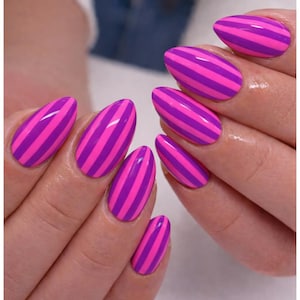 May include: A close-up of hands with almond-shaped nails. Each nail is painted with a vibrant design of alternating pink and purple stripes. The nails are glossy and well-manicured, showcasing a bold and colorful nail art style.