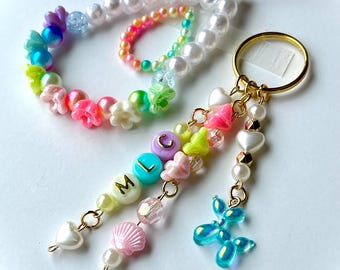 May include: A colorful beaded keychain with a gold ring. The keychain features a variety of beads, including pearls, flower-shaped beads, and beads spelling out the letters "M L C". A blue balloon dog charm and heart-shaped beads dangle from the keychain.