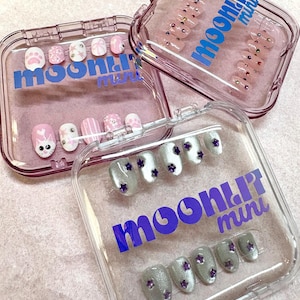 May include: Three clear plastic cases labelled moonlit mini containing sets of press-on nails. One case features pink and white nails with cat, paw, and stripe designs. Another case holds pink nails with rhinestones. The third case has silver nails with purple stars.