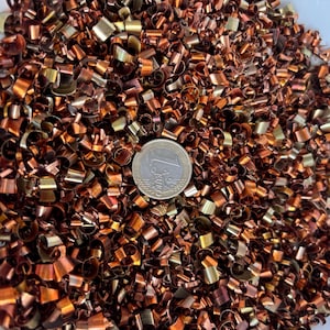 May include: A large collection of small, curled copper metal shavings with a metallic sheen. A silver and gold-colored one euro coin is placed in the center of the pile to provide a sense of scale for the tiny, twisted metal fragments.