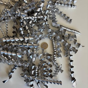 Può includere: Un mucchio di trucioli di metallo argentato, a forma di spirali, su una superficie bianca. Una moneta da euro è visibile tra i trucioli. I trucioli hanno un aspetto lucido e riflettente.