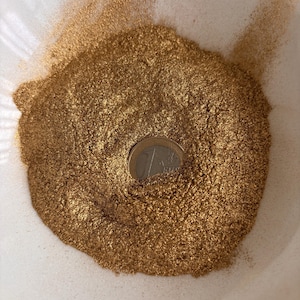 Pode incluir: Um monte de pó dourado metálico fino com uma textura cintilante e reflexiva. Uma moeda de um euro de cor prata e ouro está parcialmente inserida no centro do pó, fornecendo uma escala clara para o grão fino do material.