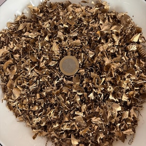 May include: A close-up shot of a pile of golden-colored metal shavings, possibly brass, filling a white bowl. A Euro coin rests on top of the shavings, providing a size reference. The shavings have a curly, textured appearance.