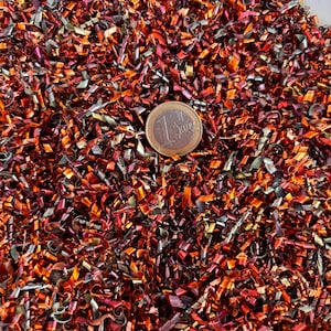 May include: A close-up shot of a pile of colorful metal shavings, predominantly in shades of red, orange, and gold. A one Euro coin is visible, providing a size reference. The metallic texture suggests a craft or industrial theme.