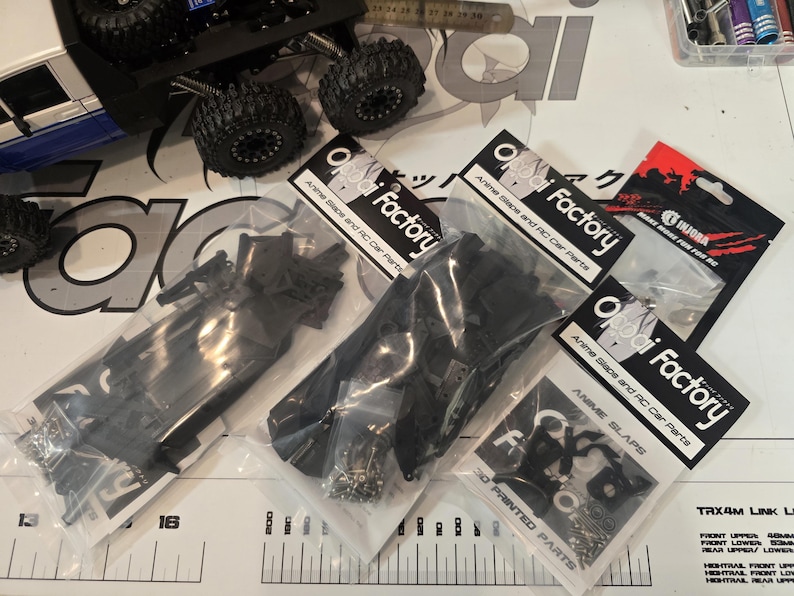 以下が含まれることがあります： Oppai FactoryとINJORAのコンポーネントを含む、RCカーパーツのいくつかのパッケージが表示されています。パッケージには黒いプラスチック部品とハードウェアが含まれています。「Anime Slaps」と「3D Printed Parts」と書かれています。RCカーとタイヤも表示されています。