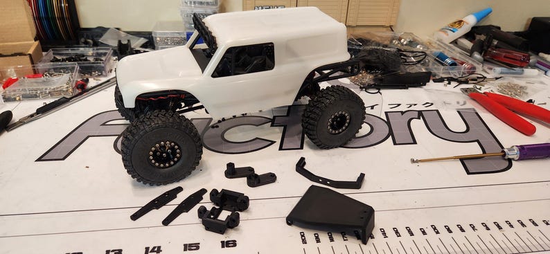 Puede incluir: Un coche de control remoto blanco todoterreno con techo negro y ruedas negras. El coche est&aacute; sobre una mesa blanca con una regla y varias piezas de pl&aacute;stico negro.