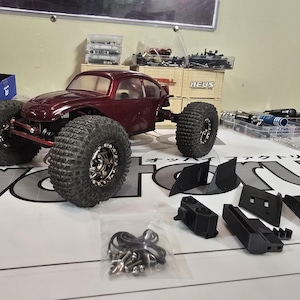 May include: A burgundy-colored remote control car with large, knobby tires. The car is a model of a classic car. Various black plastic parts and a bag of screws are arranged nearby, suggesting a hobby or model-building theme.