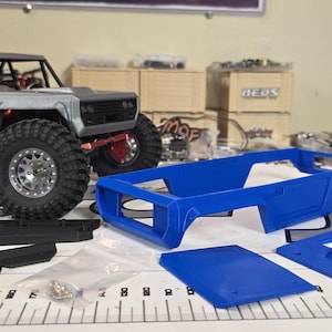 May include: A gray and black remote control truck with large tires, a roll cage, and a spare tire on top. Blue plastic components and hardware are arranged nearby, suggesting a model kit or parts for customization.