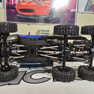 以下が含まれることがあります： 黒と青のリモコンカーシャーシ。6つの大きな黒いオフロードタイヤが付いています。シャーシは複雑なサスペンションコンポーネントを備えています。ポスターや工具を背景に、趣味のワークスペースを連想させます。