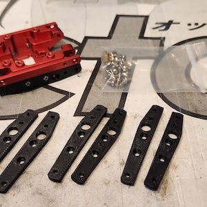May include: A selection of mechanical components, featuring a red metal part, six black elongated pieces with holes, and a small bag of silver screws. The parts are arranged on a patterned surface.