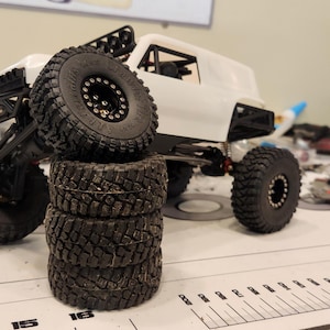 Puede incluir: Un coche RC todoterreno blanco con neum&aacute;ticos negros y una pila de neum&aacute;ticos negros y blancos. El coche est&aacute; sobre una superficie blanca con una regla.