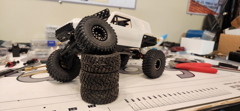 Puede incluir: Un coche RC todoterreno blanco con neum&aacute;ticos negros y una pila de neum&aacute;ticos negros y blancos. El coche est&aacute; sobre una superficie blanca con una regla.