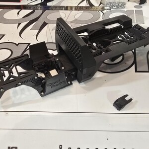 以下が含まれることがあります： 黒色の未組立モデルカーシャーシ。フレーム、サスペンションパーツなど、様々なコンポーネントが含まれています。パーツは、グレーと黒のデザインが施された白い表面に配置されています。小さな別パーツも含まれています。