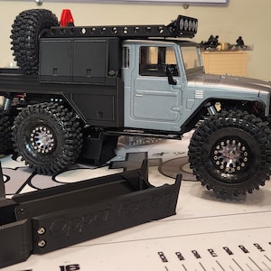 May include: A detailed model of a six-wheel-drive off-road truck, featuring a gray and black color scheme. The truck has large, knobby tires, a roof rack, and a spare tire mounted on the back. A black component is in the foreground.
