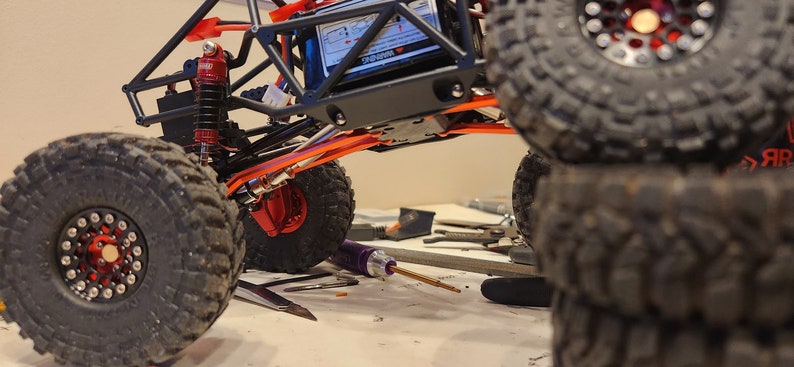 K&ouml;nnte beinhalten: Ein schwarzer und orangefarbener ferngesteuerter Rock-Crawler mit gro&szlig;en schwarzen Reifen. Das Auto ist angehoben und die Reifen sind &uuml;bereinander gestapelt. Das Auto hat ein schwarzes Chassis und einen roten Sto&szlig;d&auml;mpfer.