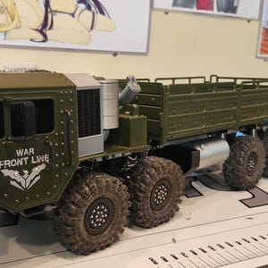 Pu&ograve; includere: Un modello di camion militare verde con un pianale e una cabina con un parabrezza. Il camion ha un logo "WAR FRONT LINE" sul lato. Il camion &egrave; seduto su un tavolo con un righello.