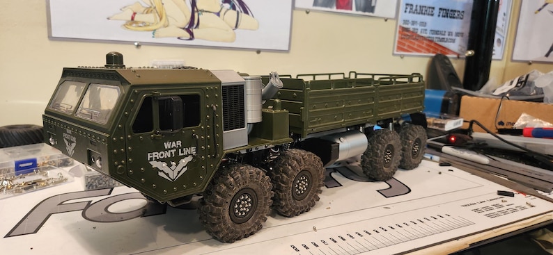 Pu&ograve; includere: Un modello di camion militare verde con un pianale e una cabina con un parabrezza. Il camion ha un logo "WAR FRONT LINE" sul lato. Il camion &egrave; seduto su un tavolo con un righello.