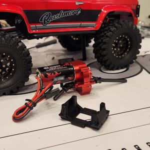 May include: A red, black, and silver toy truck with a red and black motor and a black plastic mount. The truck has the word "Rustler" printed on the side.