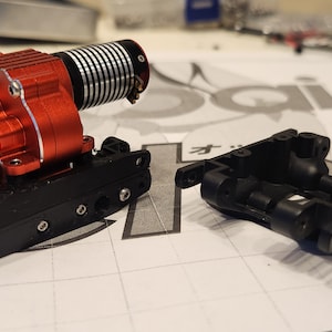 K&ouml;nnte beinhalten: Ein roter und schwarzer Motor mit einem schwarzen Getriebe und einem schwarzen Chassis. Der Motor ist mit Schrauben am Chassis befestigt. Das Chassis besteht aus Kunststoff und hat ein Gittermuster.