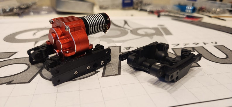 K&ouml;nnte beinhalten: Ein roter und schwarzer Motor mit einem schwarzen Getriebe und einem schwarzen Chassis. Der Motor ist mit Schrauben am Chassis befestigt. Das Chassis besteht aus Kunststoff und hat ein Gittermuster.