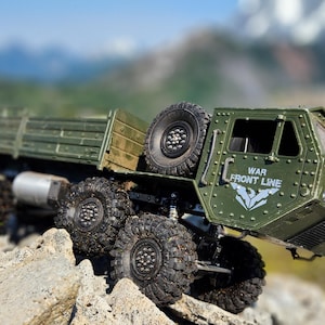 Pu&ograve; includere: Un camion militare giocattolo verde con un cassone e le parole "WAR FRONT LINE" sul lato. Il camion &egrave; su una superficie rocciosa con una catena montuosa sullo sfondo.