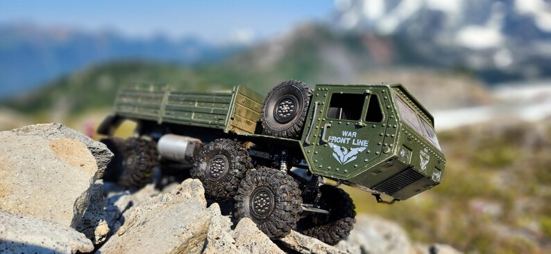 Pu&ograve; includere: Un camion militare giocattolo verde con un cassone e le parole "WAR FRONT LINE" sul lato. Il camion &egrave; su una superficie rocciosa con una catena montuosa sullo sfondo.