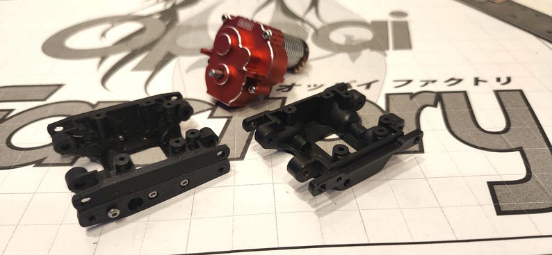K&ouml;nnte beinhalten: Zwei schwarze Kunststoffteile f&uuml;r ein Modellfahrzeug. Die Teile sind so konzipiert, dass sie einen Motor oder andere Komponenten aufnehmen k&ouml;nnen. Die Teile befinden sich auf einer wei&szlig;en Oberfl&auml;che mit einem Gittermuster.