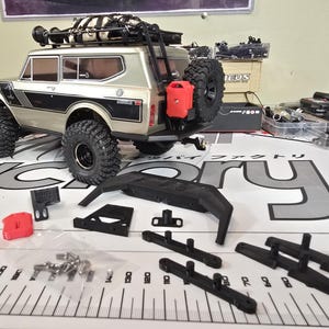 May include: A detailed image of a model RC truck, silver and black, with large off-road tires. Accessories include a spare tire, a red fuel canister, and various black components. The truck is on a surface with a ruler and other parts.