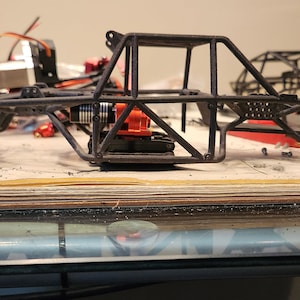 K&ouml;nnte beinhalten: Ein schwarzer Metallrahmen f&uuml;r ein ferngesteuertes Auto mit einem orangefarbenen Motor und einem blauen Schraubendreher auf einem Holztisch.