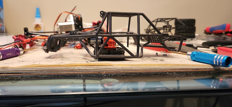 K&ouml;nnte beinhalten: Ein schwarzer Metallrahmen f&uuml;r ein ferngesteuertes Auto mit einem orangefarbenen Motor und einem blauen Schraubendreher auf einem Holztisch.