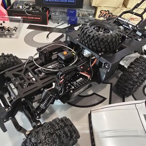 以下が含まれることがあります： 黒色のRCカーシャーシ。 大きなテクスチャードタイヤとフレームに取り付けられたスペアタイヤが特徴です。 シャーシには、バッテリーや制御モジュールなど、さまざまな電子コンポーネントが含まれています。 画像は部分的に組み立てられたモデルを示しています。
