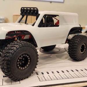 Puede incluir: Un coche RC todoterreno blanco, personalizado, con ruedas negras y una jaula antivuelco negra. El coche tiene una carrocer&iacute;a blanca con un techo negro y una luneta trasera negra. El coche est&aacute; sobre una superficie blanca.
