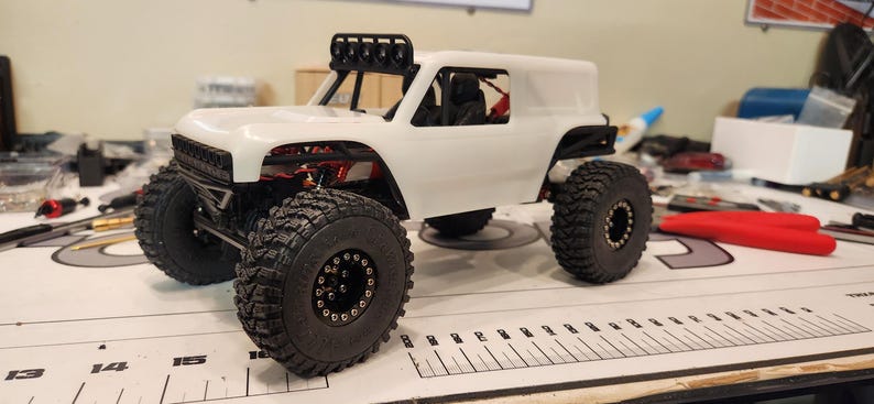 Puede incluir: Un coche RC todoterreno blanco, personalizado, con ruedas negras y una jaula antivuelco negra. El coche tiene una carrocer&iacute;a blanca con un techo negro y una luneta trasera negra. El coche est&aacute; sobre una superficie blanca.