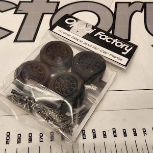 Pu&ograve; includere: Un pacchetto di quattro pneumatici in gomma nera e ferramenta per un'auto RC. La confezione &egrave; etichettata "Ai Factory" e "Alloy Wheels and RC Car Parts".