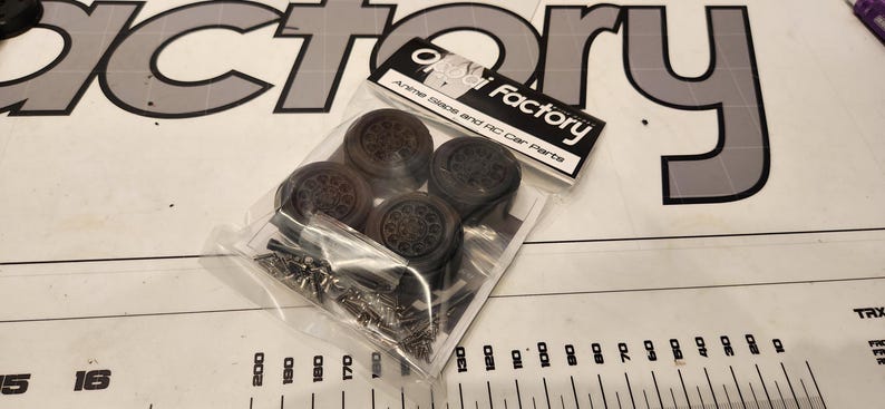 Pu&ograve; includere: Un pacchetto di quattro pneumatici in gomma nera e ferramenta per un'auto RC. La confezione &egrave; etichettata "Ai Factory" e "Alloy Wheels and RC Car Parts".