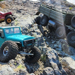 Pu&ograve; includere: Tre veicoli fuoristrada giocattolo su un terreno roccioso. Un camion blu turchese con pneumatici grandi, un camion rosso con un cassone nero e un camion militare verde con una piattaforma.
